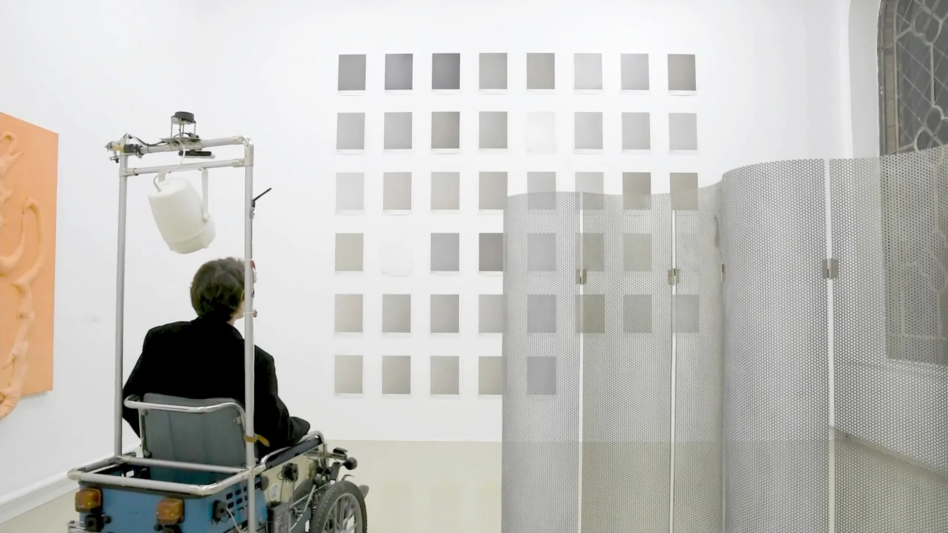 A person sits in a modified electric wheelchair with attachments in front of a wall with multiple photos