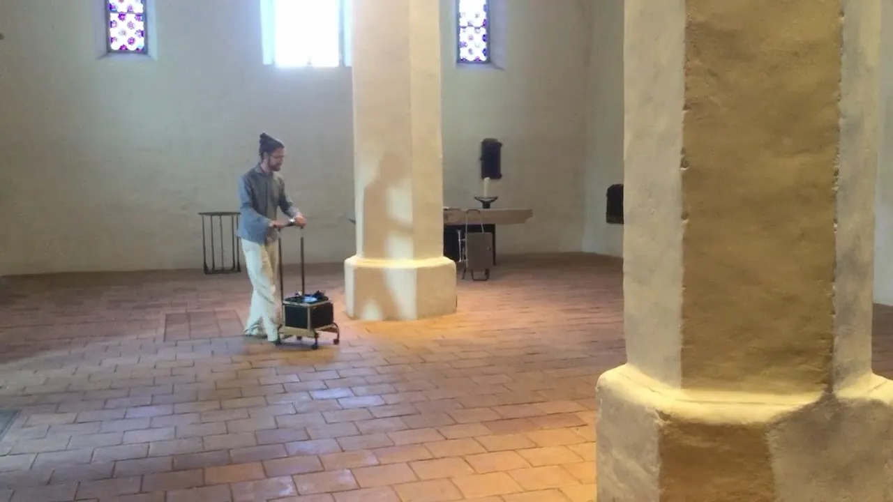 A dark chapel room, a small cart with equipment stands before the altar, a candle burns on the floor