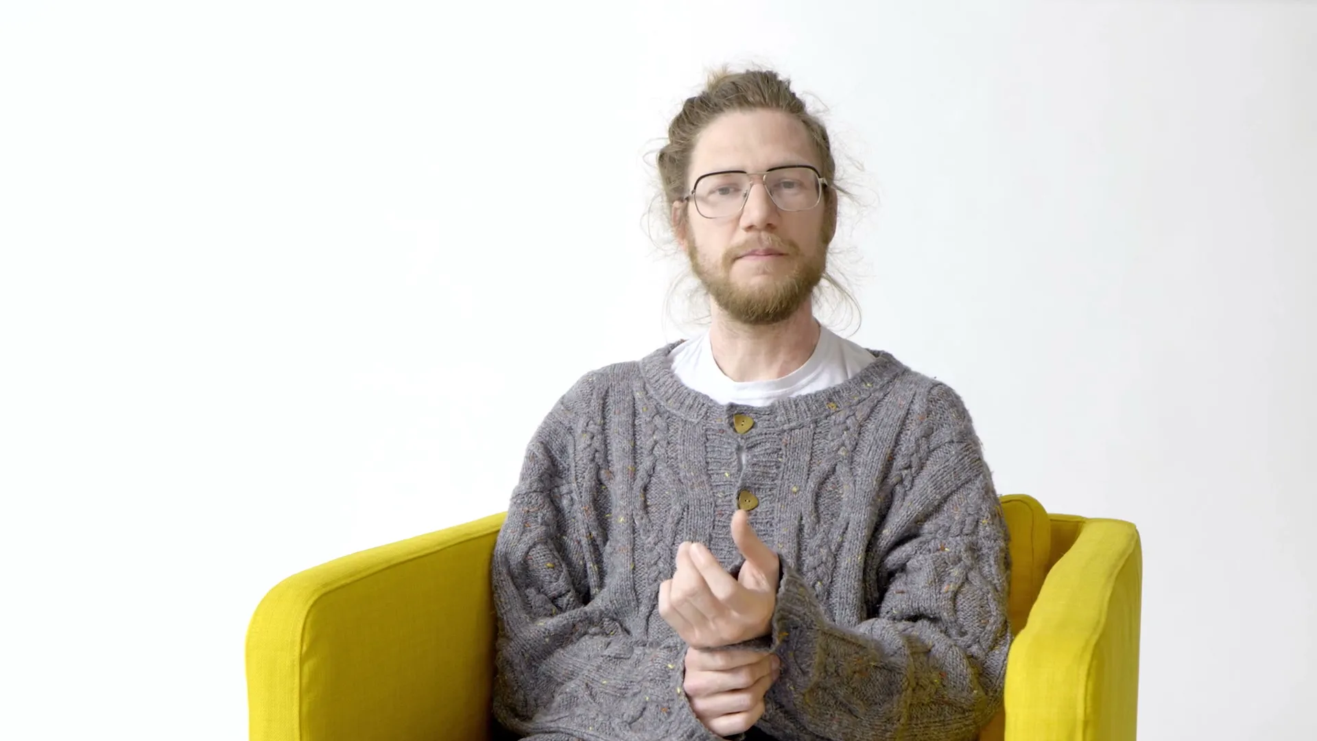 A person with glasses and tied-up hair sits in a yellow armchair in front of a white wall, speaking to the camera